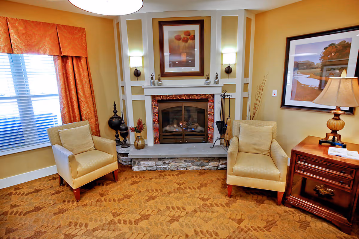 A cozy living room area with two beige armchairs facing a stone fireplace with a fire burning inside. The fireplace is framed with white wood and has a decorative picture above it. To the right of the fireplace is a wooden side table with a lamp and a framed landscape picture on the wall. To the left is a window with orange patterned curtains and white blinds. The floor is carpeted with a patterned design in warm tones.
