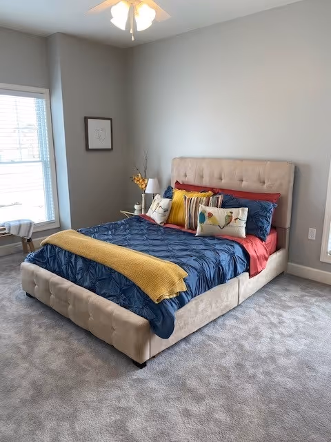 A neatly made bed with a beige upholstered headboard and base, covered with a dark blue quilt, red sheets, and multiple decorative pillows in various colors including yellow and white with bird designs. A yellow throw blanket is draped across the foot of the bed. The room has light gray walls, a ceiling fan with lights, a window with blinds, a small side table with a lamp and decorative items, and light gray carpeted flooring.