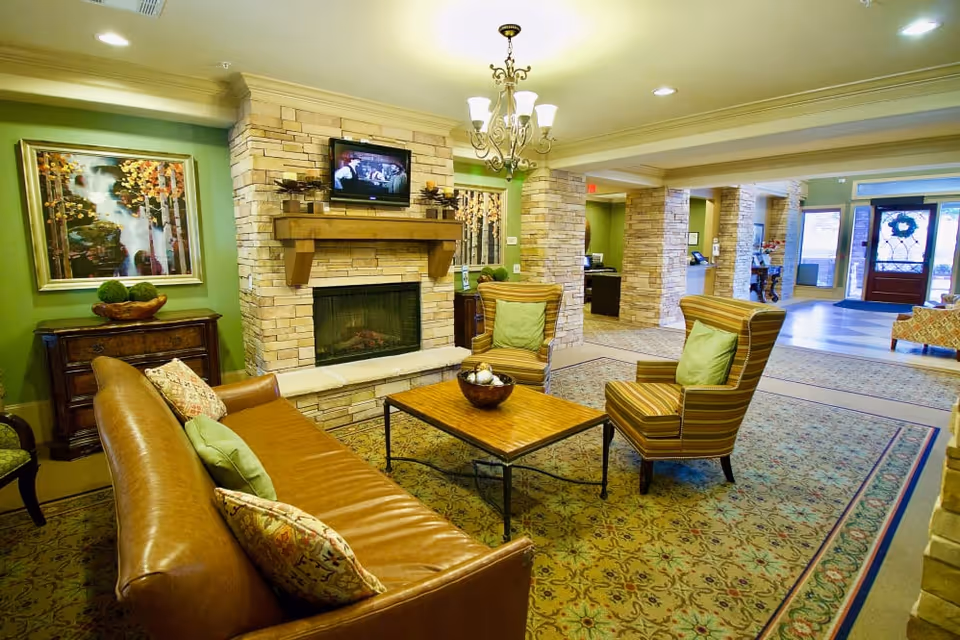 A cozy senior living facility common area with a brown leather sofa, two striped armchairs with green pillows, a wooden coffee table, and a stone fireplace with a mounted TV above it. The walls are painted green and decorated with framed artwork. The room has a patterned carpet and a chandelier hanging from the ceiling. In the background, there is a reception desk and an entrance door with a wreath.