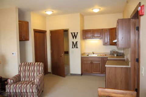 Interior view of a living area in an assisted living facility featuring a patterned armchair, a small kitchen with wooden cabinets, a countertop, and a sink. There are two open doors leading to other rooms, and the letters 'W' and 'M' are mounted on the wall between the doors. The walls are painted beige and the floor is carpeted.