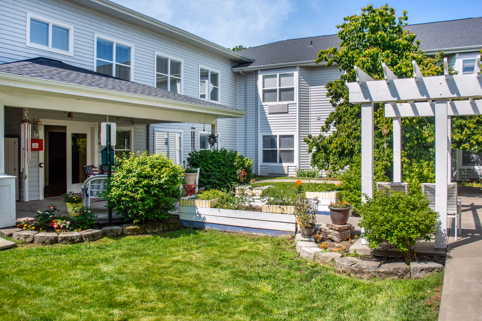 Outdoor courtyard area of a senior living facility with green grass, bushes, flowers, and a white pergola. The building has light gray siding with multiple windows and a covered entrance area.