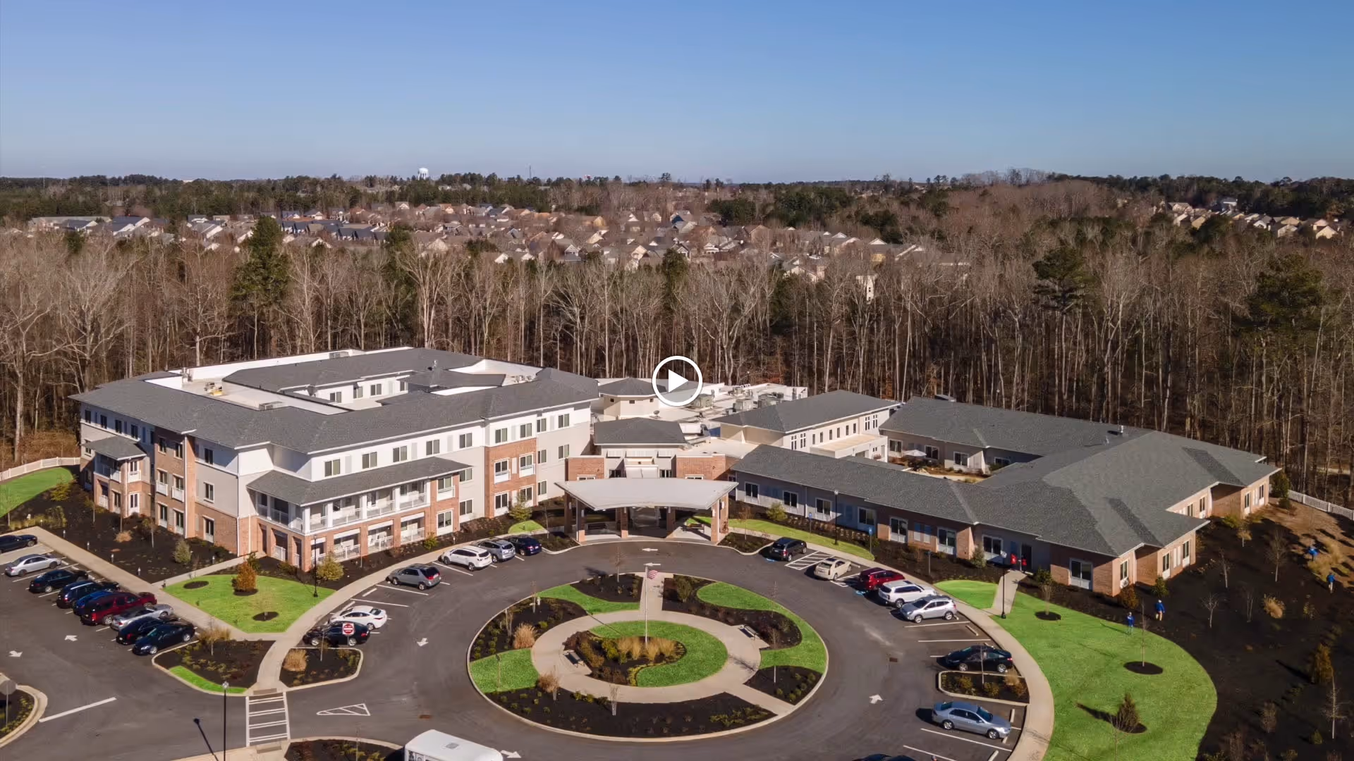 Aerial view of Westhill Newnan Crossing senior living facility showing a large, multi-wing building with a circular driveway and parking area surrounded by green lawns and trees, with a wooded area and residential neighborhood in the background under a clear blue sky.