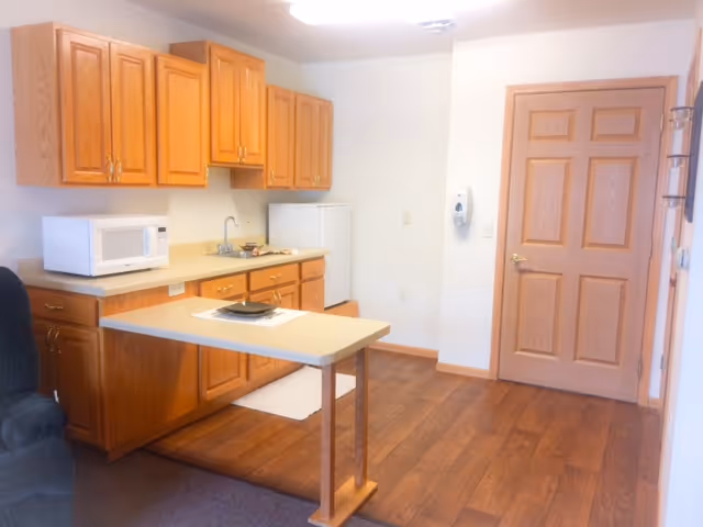 Small kitchenette with wooden cabinets, microwave, mini fridge, sink, and a peninsula table in a senior living unit.