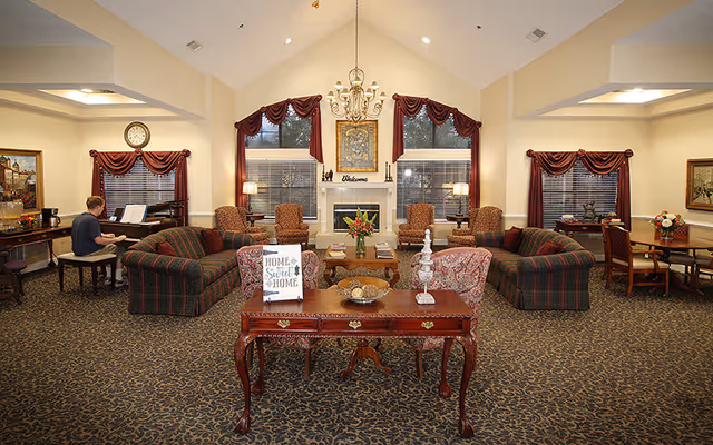 A spacious living room area in a senior living facility with high vaulted ceilings, patterned carpet, and multiple seating arrangements including sofas and armchairs. A person is playing the piano on the left side. There is a fireplace in the center with a 'Welcome' sign above it, flanked by two large windows with red curtains. A wooden table with decorative items and a 'Home Sweet Home' sign is in the foreground.