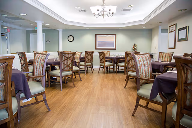 A dining room with wood floors, several tables set with dark tablecloths and wooden chairs under a chandelier.