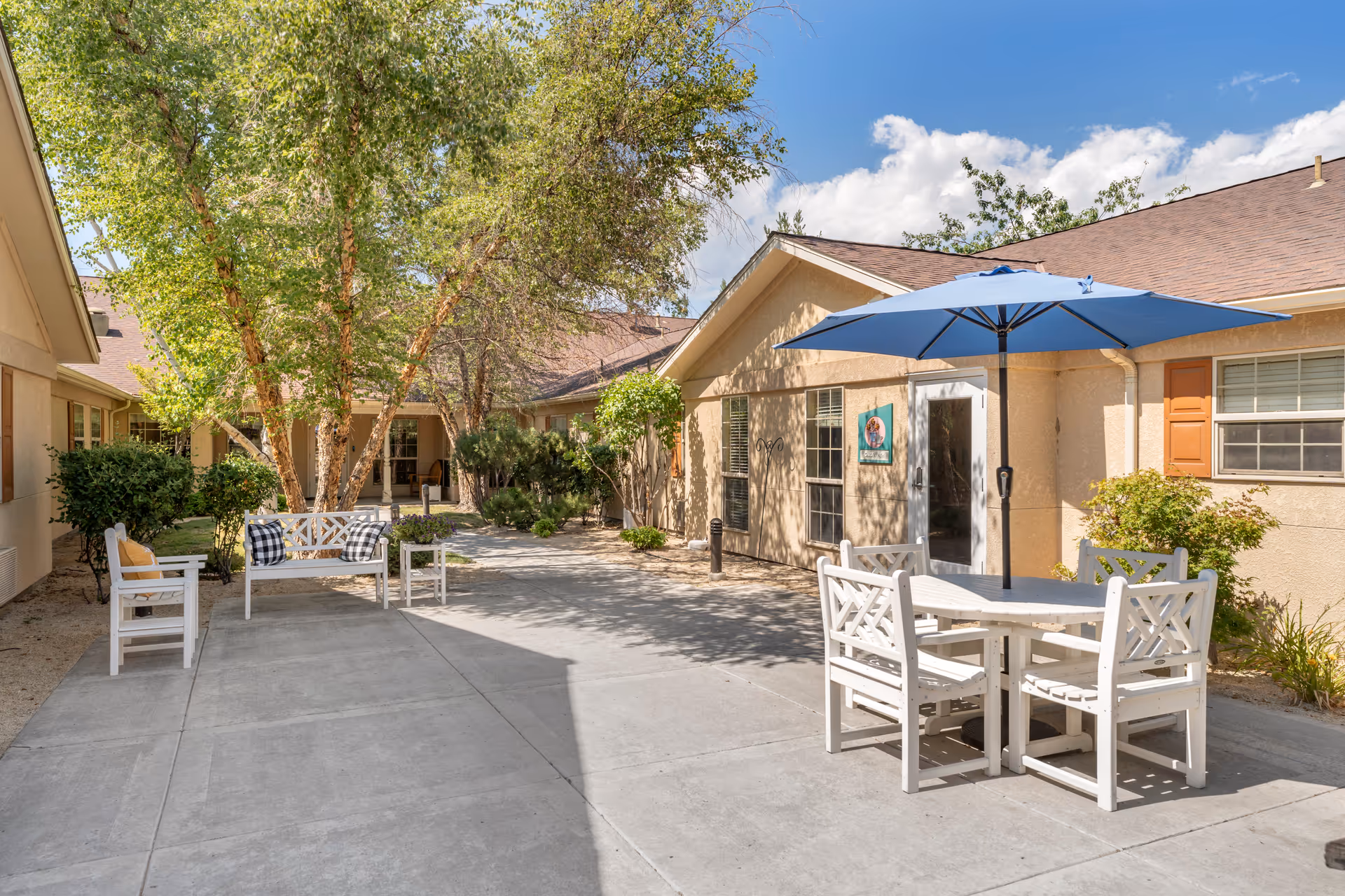Outdoor courtyard area at Brookdale Reno featuring a concrete walkway, white wooden benches with cushions, a white wooden table with four chairs under a blue umbrella, surrounded by trees and shrubs, with beige building walls and windows in the background under a blue sky with some clouds.