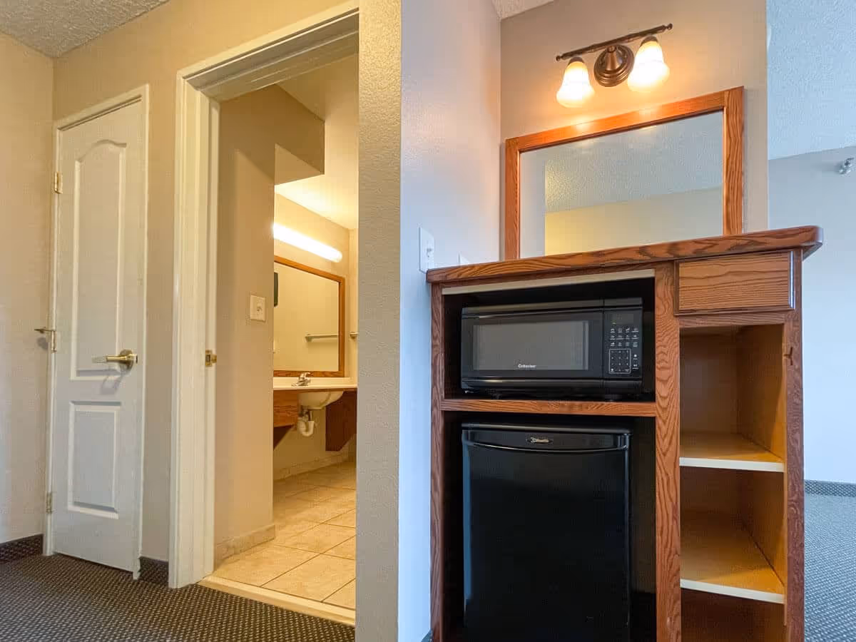 Interior view of a room showing a wooden cabinet with a microwave and mini-fridge next to an open doorway revealing a bathroom sink and mirror.
