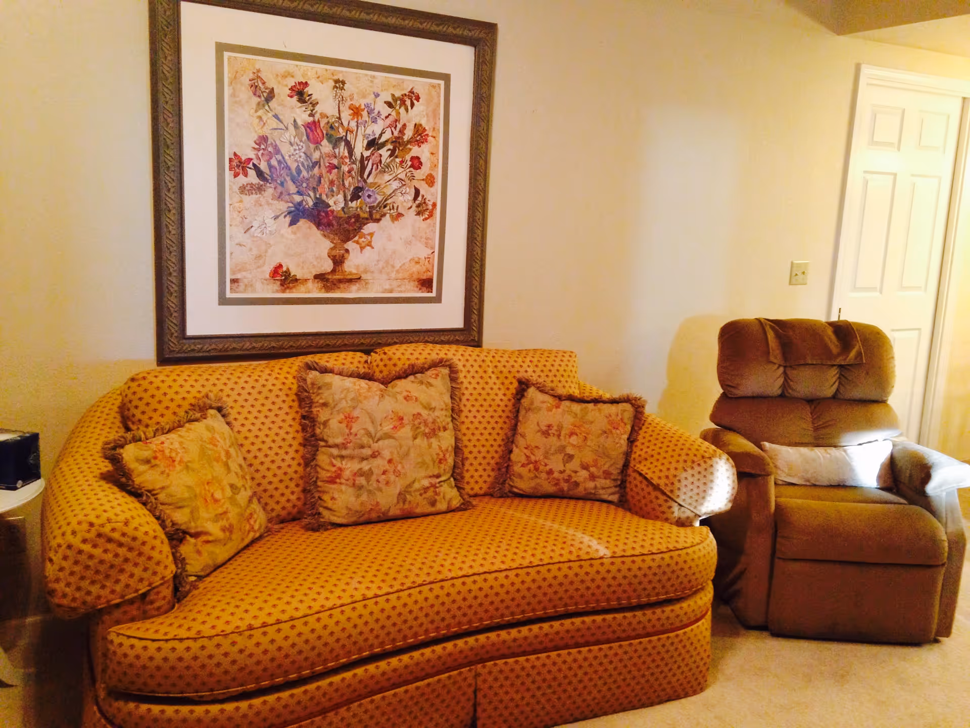 A cozy living room area with a patterned yellow sofa adorned with three floral cushions and a brown recliner chair next to it. Above the sofa hangs a framed painting of a floral arrangement. The walls are light-colored, and a white door is visible in the background.