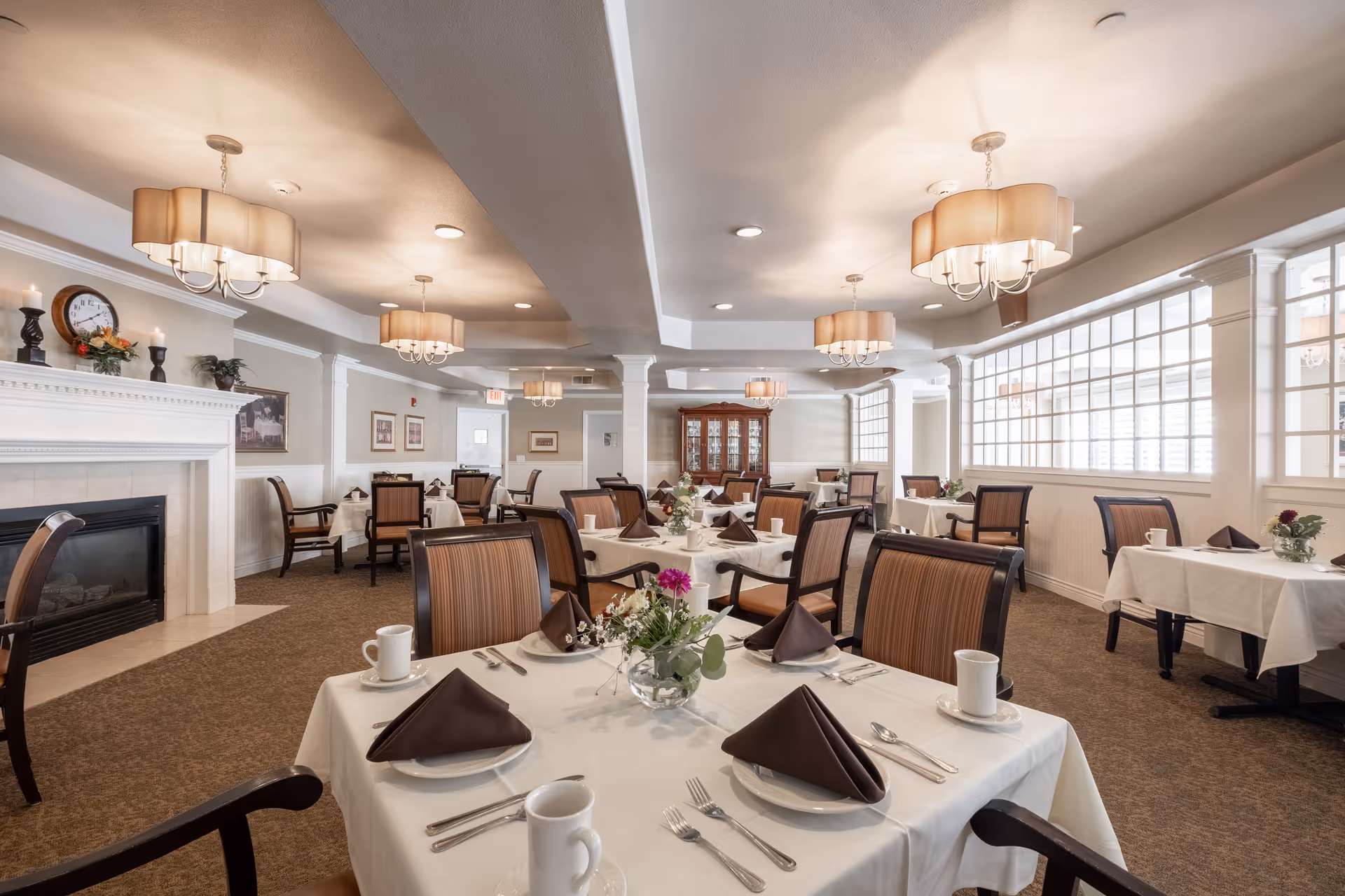 A spacious dining room in a senior living facility with multiple tables covered in white tablecloths, each set with brown folded napkins, white plates, silverware, and white coffee mugs. The room features beige walls, a carpeted floor, large windows with white framing, elegant chandeliers, a fireplace with candles and a clock on the mantel, and a wooden china cabinet in the background.