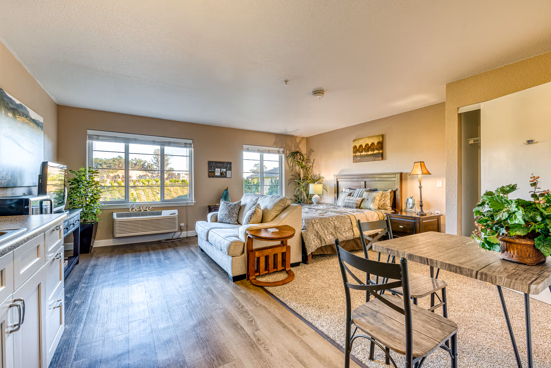 A bright and cozy studio apartment in a senior living facility featuring a bed with pillows and a blanket, a beige sofa with cushions, a small wooden side table, a dining table with two chairs, a kitchenette with white cabinets and a sink, and large windows letting in natural light. The room is decorated with plants, a lamp, and wall art.