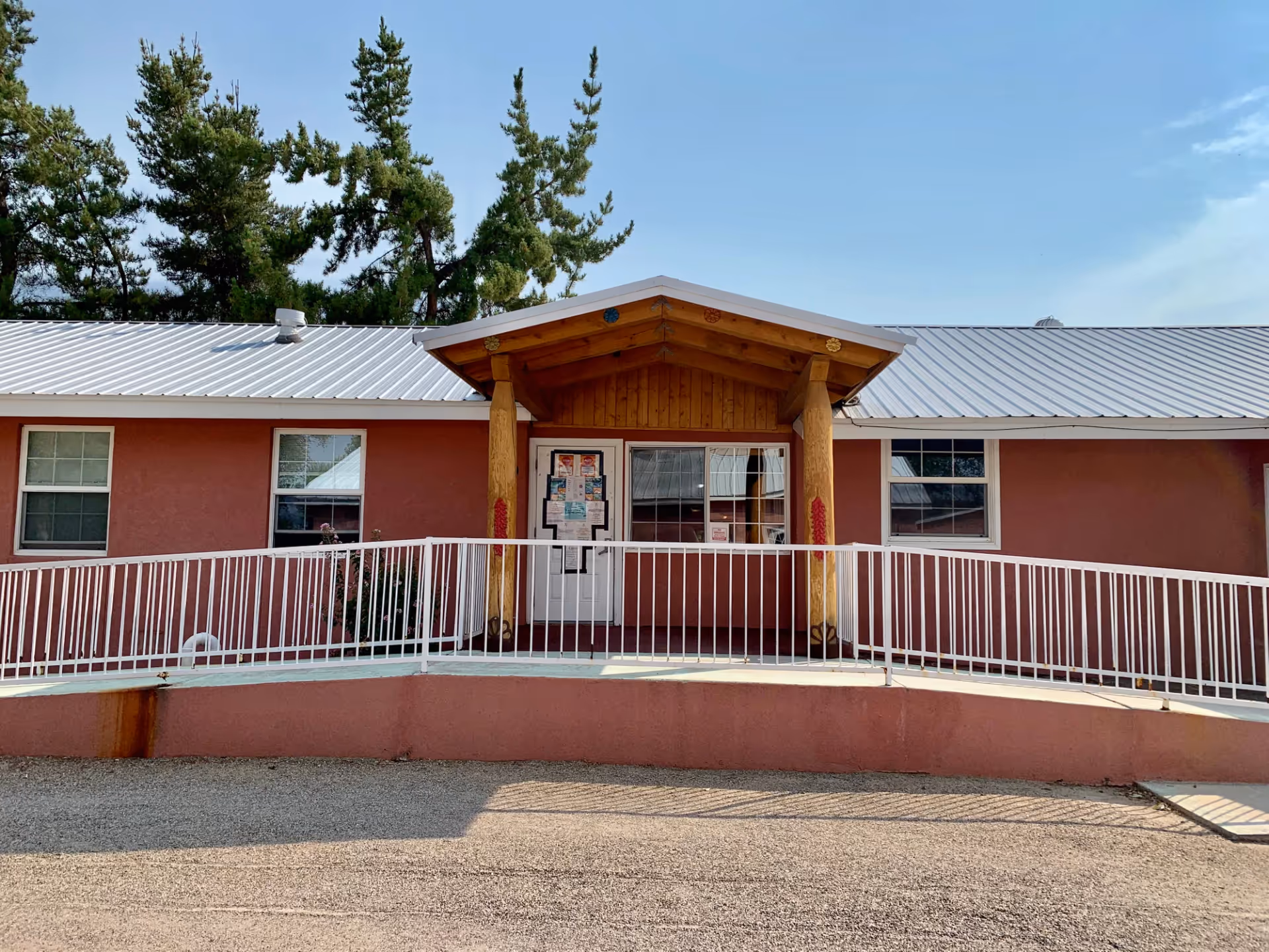 Front exterior view of Casa de Rosa Assisted Living facility with a pinkish exterior wall, a metal roof, a covered entrance supported by wooden beams, and a white ramp railing leading to the door. There are two windows on either side of the entrance and trees in the background under a clear blue sky.