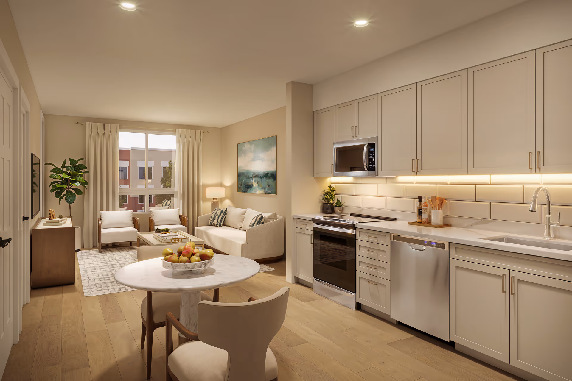 A modern open-concept living space featuring a kitchen and living room. The kitchen has white cabinets, a stainless steel oven, microwave, dishwasher, and a sink with a faucet. Under-cabinet lighting illuminates the white subway tile backsplash. The living room includes a beige sofa with cushions, two armchairs, a coffee table, a side table with a lamp, and a large window with curtains letting in natural light. A round dining table with chairs and a bowl of fruit is in the foreground. Light wood flooring runs throughout the space.