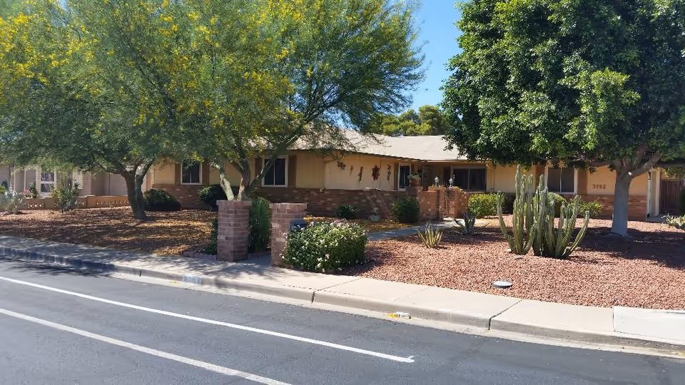 Single-story beige senior care home exterior with desert landscaping and large trees.