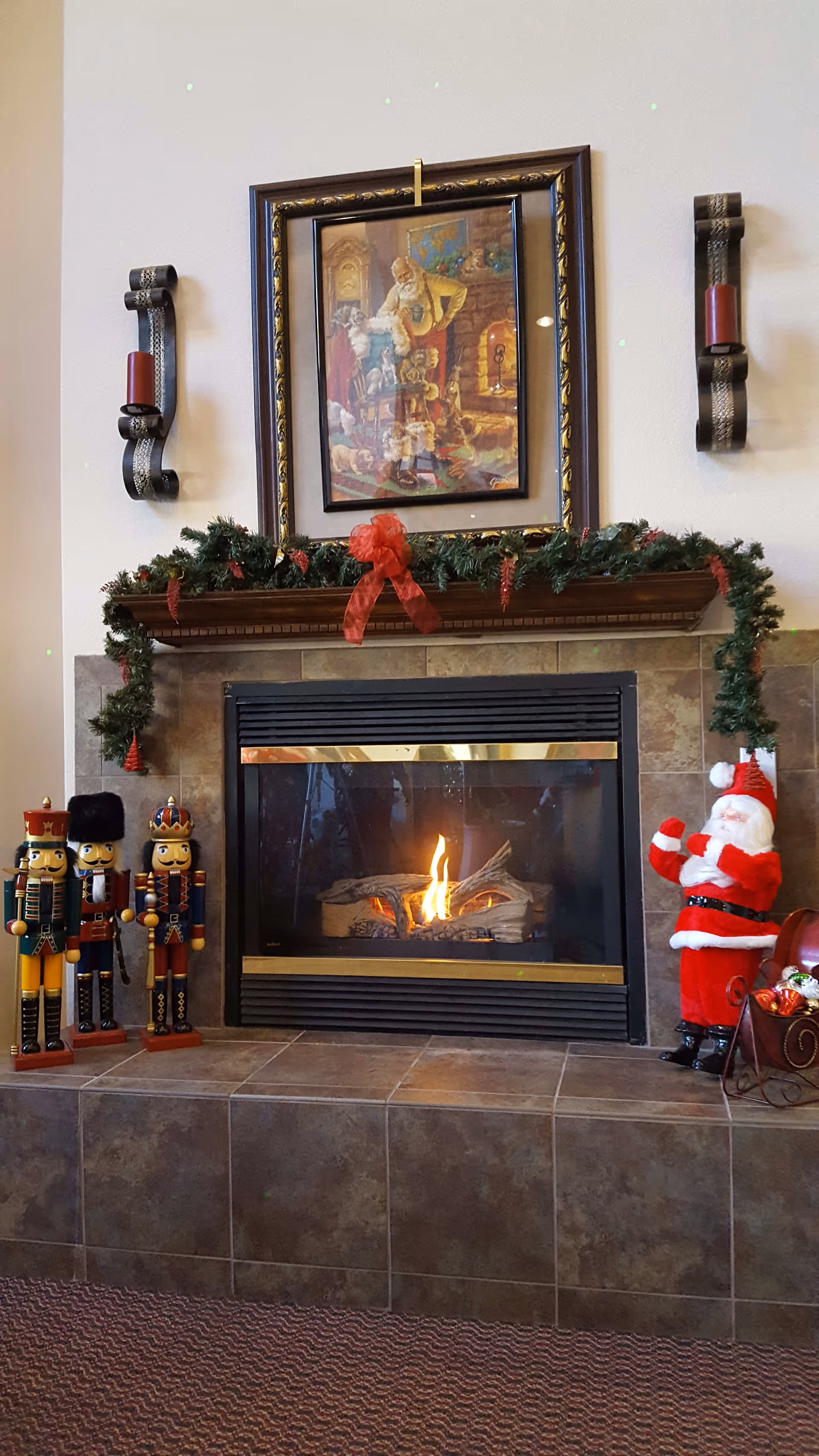 A cozy fireplace decorated for Christmas with a garland and a red bow on the mantel. Above the mantel is a framed painting depicting a man with dogs near a fireplace. On the left side of the fireplace are three nutcracker figurines, and on the right side is a Santa Claus figure next to a small sleigh filled with ornaments.