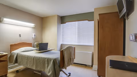 A simple, clean bedroom in a healthcare facility with a single bed covered in a green bedspread, a small bedside table, a laptop on a tray over the bed, a window with closed blinds, a wooden wardrobe, and a wall-mounted television.