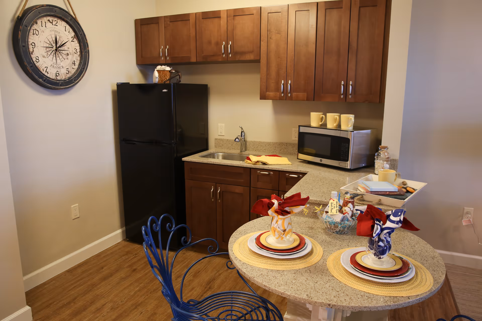A small kitchen area with wooden cabinets, a black refrigerator, a microwave, and a sink. A round countertop table is set with two place settings, each with plates, cups, and folded napkins. A large decorative wall clock is mounted on the wall.