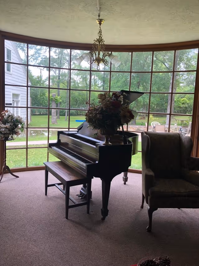 Interior living room with a grand piano and bench by a large curved window overlooking a green yard, with an armchair and floral arrangements.