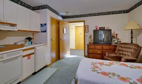 Interior view of a senior living facility room featuring a small kitchenette with white cabinets, a dishwasher, and a sink on the left side. To the right, there is a bed with a floral quilt, a brown cushioned armchair, a wooden dresser with a vintage TV on top, a table lamp, and some decorative items including flowers. The room has light-colored walls with floral wallpaper border near the ceiling and a carpeted floor. A door is open leading to a hallway with natural light.