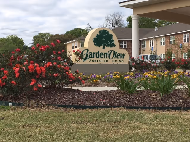 Outdoor view of the Garden View Assisted Living facility sign surrounded by colorful flowers and landscaping, with the building and parked cars visible in the background under a cloudy sky.
