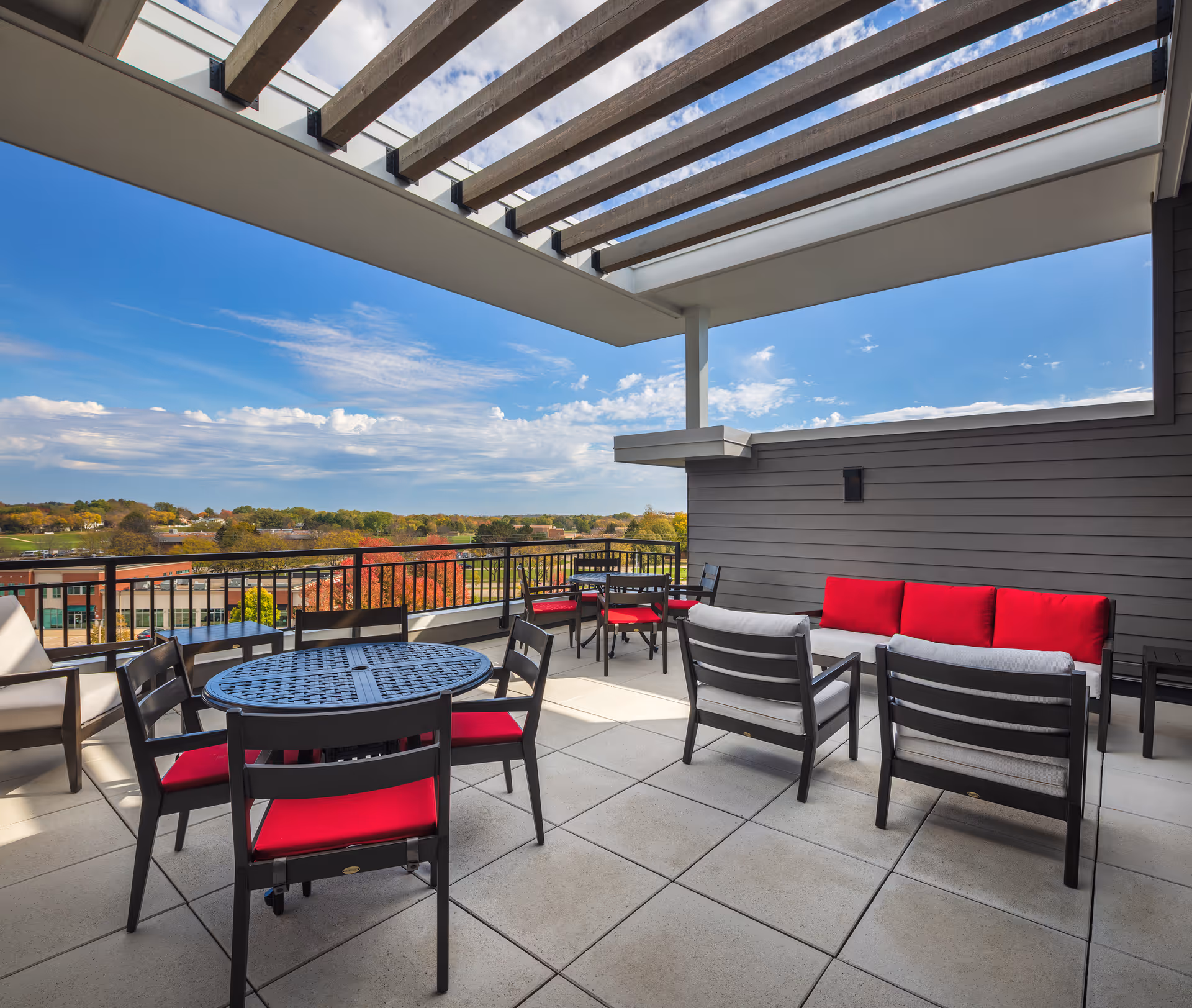 Outdoor patio area with modern black metal tables and chairs featuring red cushions. The patio has a partial pergola overhead and overlooks a scenic view of trees and buildings under a partly cloudy blue sky.