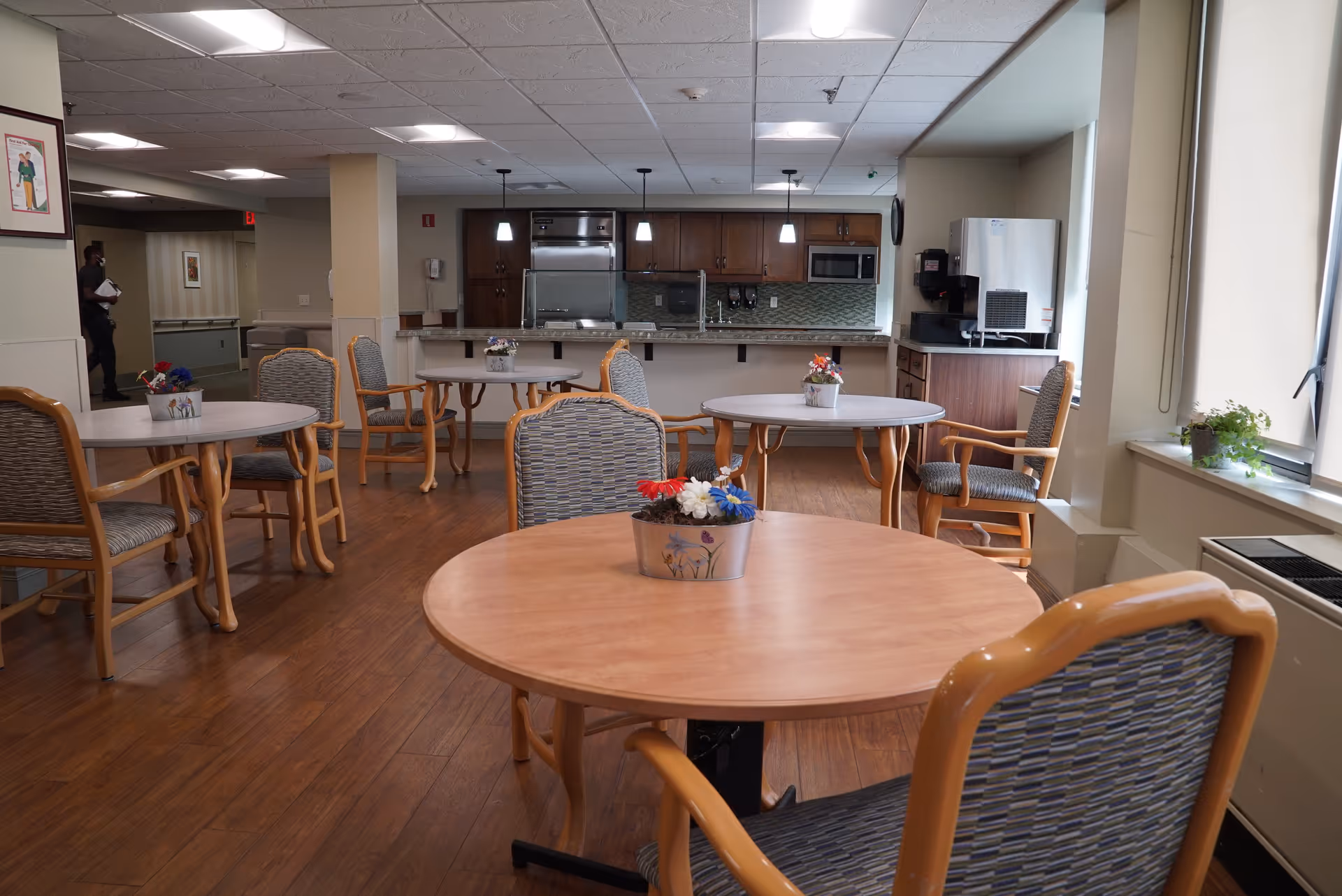 Dining room in a senior living facility with round wooden tables and chairs, small flower centerpieces, and a kitchen/service counter in the background.