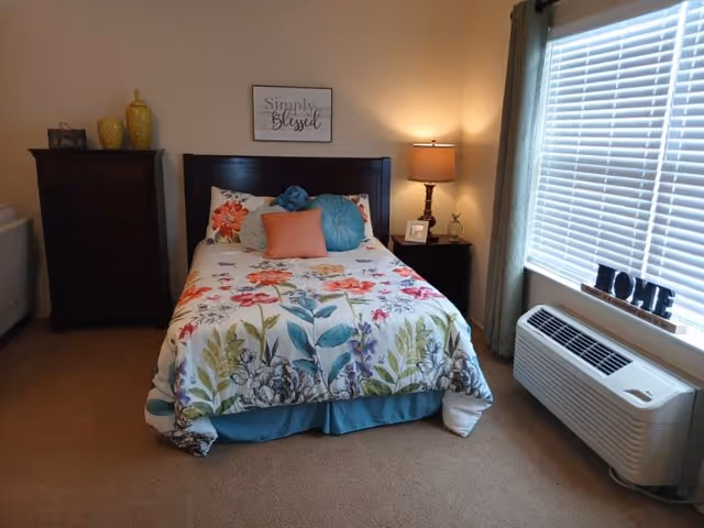 A neatly made bed with colorful floral bedding in a cozy bedroom with a dresser, nightstand, lamp and a window air conditioner.