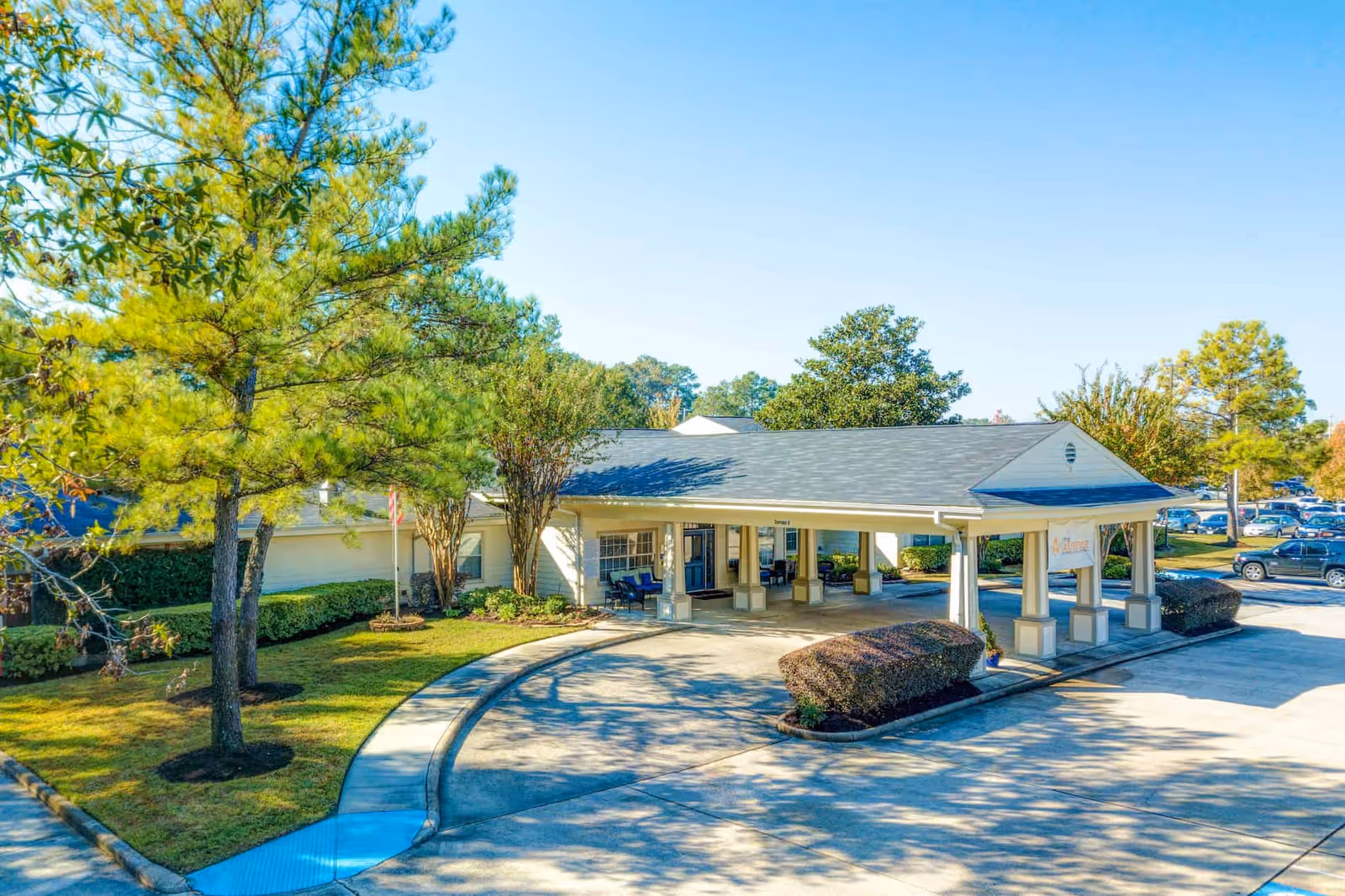 Exterior view of The Auberge at Kingwood senior living facility entrance with a covered drop-off area, surrounded by trees and landscaped bushes under a clear blue sky.