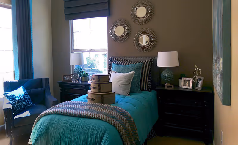 A styled bedroom with a teal-covered bed, bedside tables and lamps, decorative mirrors above the headboard, and an armchair by the window.