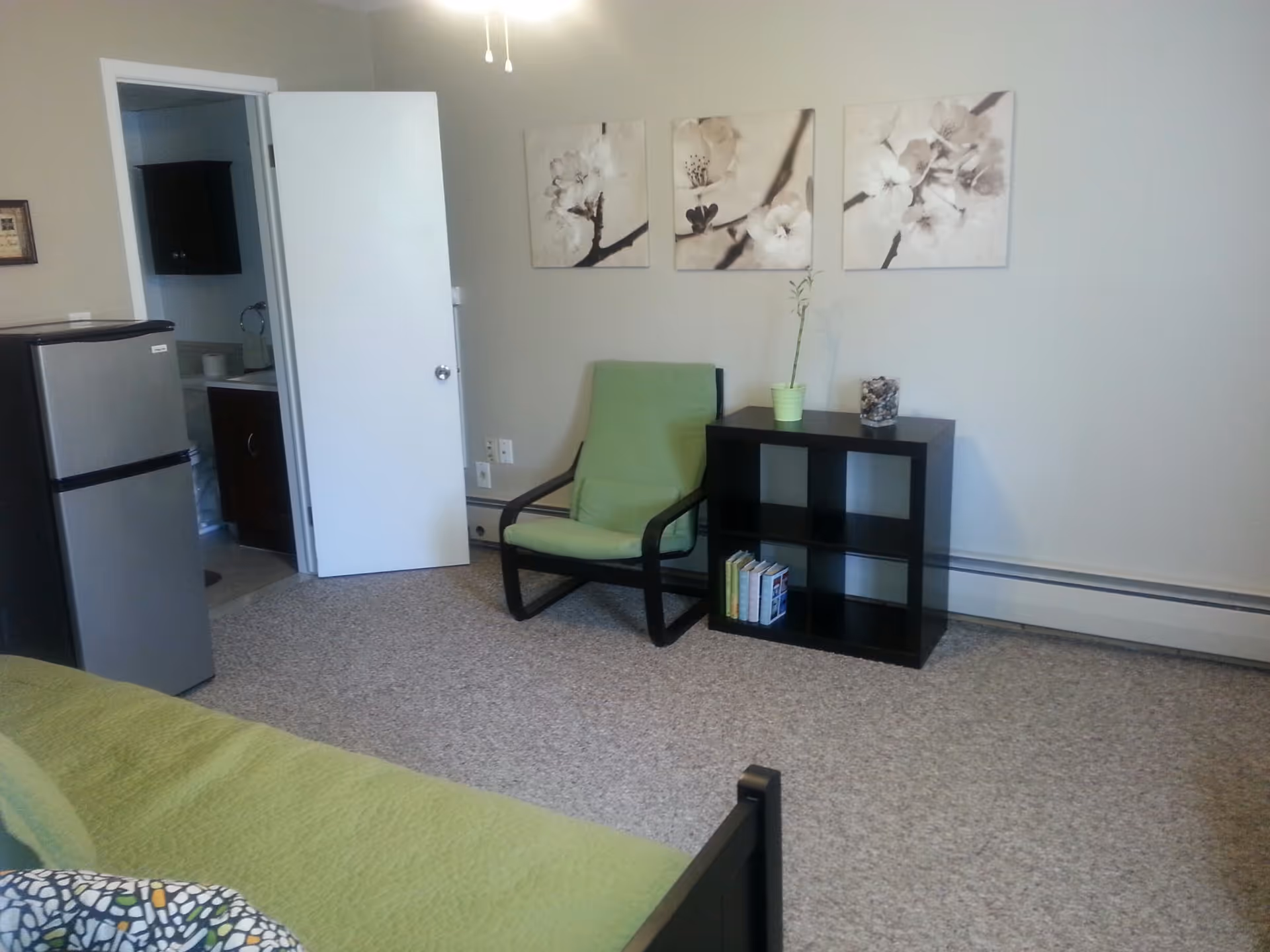 Studio bedroom with a green chair, cube shelf, mini refrigerator, and an open bathroom door.