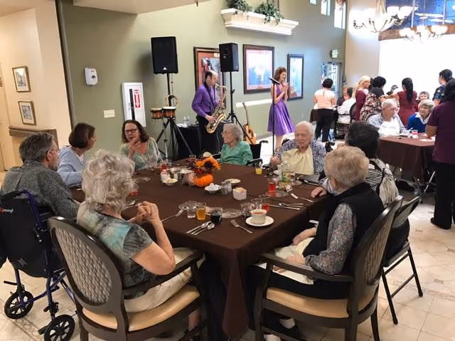 A group of elderly people seated around a large table with brown tablecloth in a dining area, enjoying drinks and conversation. In the background, a man playing a saxophone and a woman singing with a microphone entertain the guests. Other people are standing and socializing near another table. The room has light green walls, framed pictures, and a chandelier.