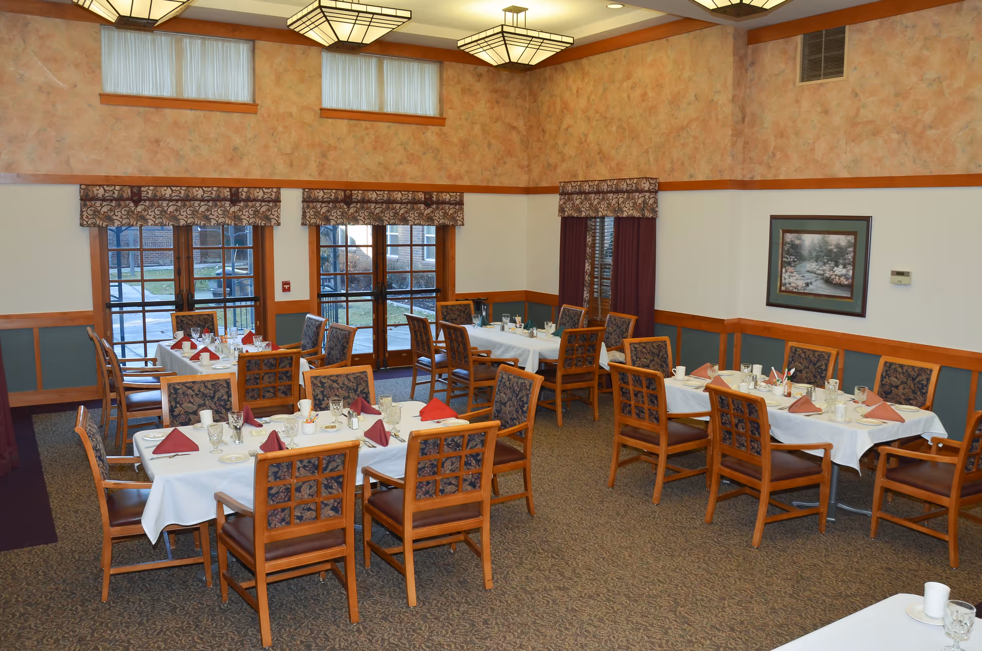 A dining room with several tables covered in white tablecloths, each set with glasses, cups, plates, and folded red and brown napkins. The room has patterned carpet, wooden chairs with upholstered backs, large windows with decorative valances, and warm-toned walls with a framed painting.