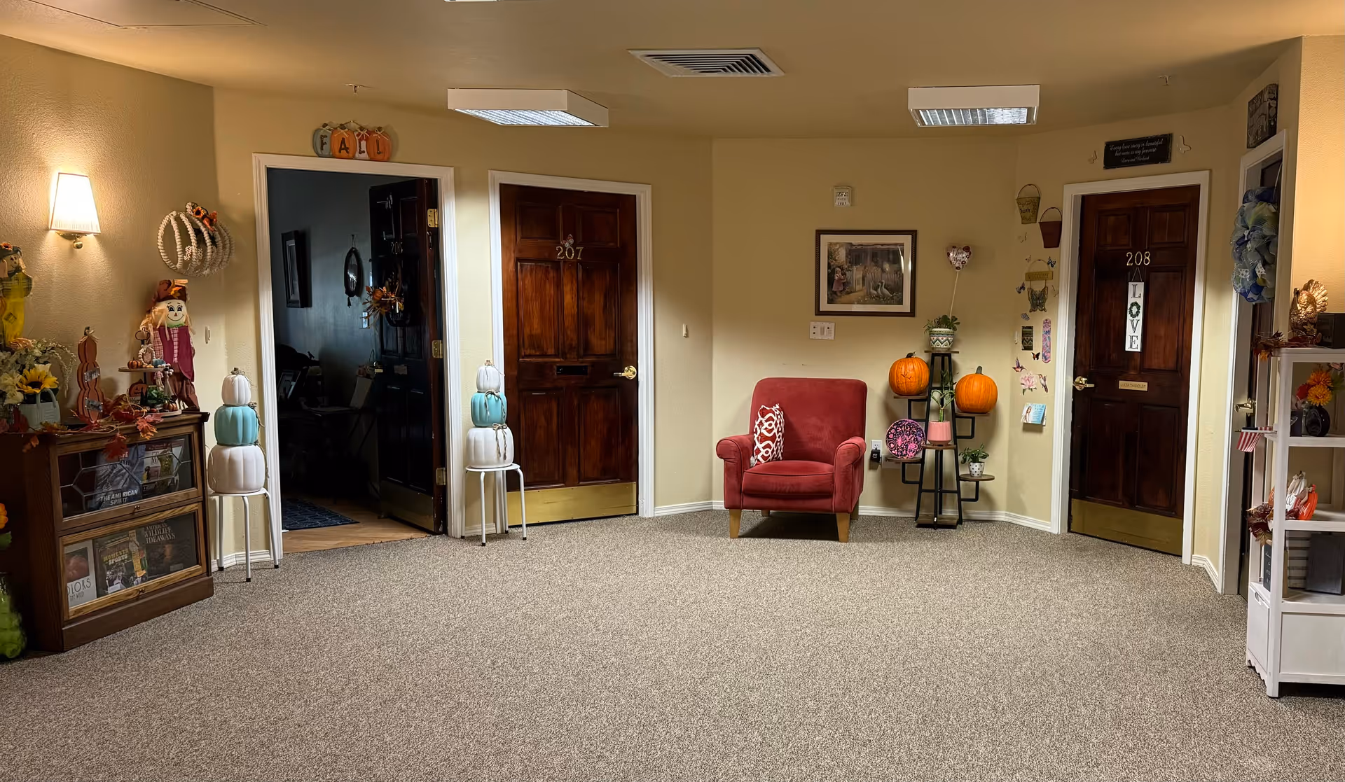 A cozy interior hallway area in a senior living facility with two wooden doors labeled 207 and 208. The space is decorated with fall-themed items including pumpkins, a scarecrow figure, and seasonal wreaths. There is a red armchair with a patterned pillow between the doors, and shelves with various decorative items on either side. The walls are painted light yellow and the floor is carpeted.
