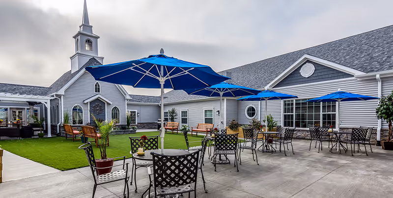 Outdoor patio area at a senior living facility with several black metal tables and chairs, each table shaded by a blue umbrella. The patio is adjacent to a building with gray siding and white trim, featuring large windows and a small steeple. There are potted plants and benches around the grassy courtyard area.