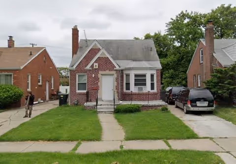 Small brick house with white door, front steps, green lawn, and parked cars.