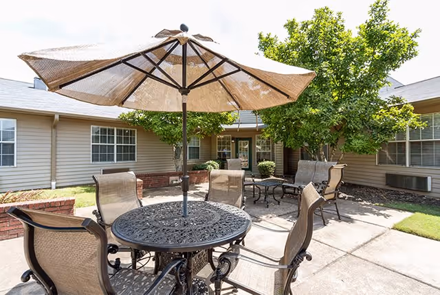 Outdoor patio area at Shelby Gardens Place with a round metal table and four mesh chairs under a large beige umbrella. Additional seating with cushioned chairs and a small table is visible in the background, surrounded by trees and the exterior walls of the building.