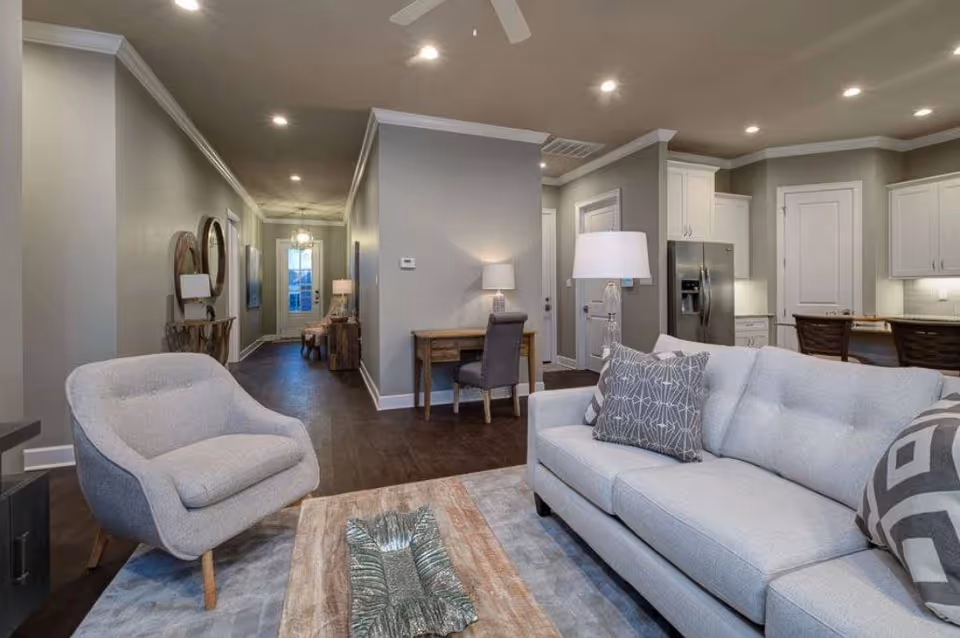 A modern living room with a light gray sofa adorned with patterned pillows, a matching armchair, a wooden coffee table with a decorative centerpiece, and a lamp on a side table. The room opens into a kitchen area with white cabinets, stainless steel refrigerator, and a breakfast bar with stools. The space features recessed lighting, a ceiling fan, and a hallway leading to a door with glass panels.