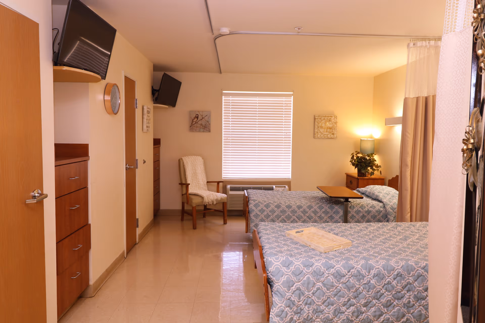 A well-lit room with two single beds covered in blue patterned bedspreads. There is a wooden chair with a white blanket draped over it near a window with closed blinds. A small wooden nightstand with a lamp and a plant is positioned between the beds. Two wall-mounted televisions are visible, one near the ceiling on the left wall and another on the far wall. The room has light-colored walls and a tiled floor, with a door and a set of drawers on the left side.