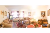 Bright communal living room with sofas, a coffee table on a patterned rug, a TV, and wall artwork beneath curtained windows.