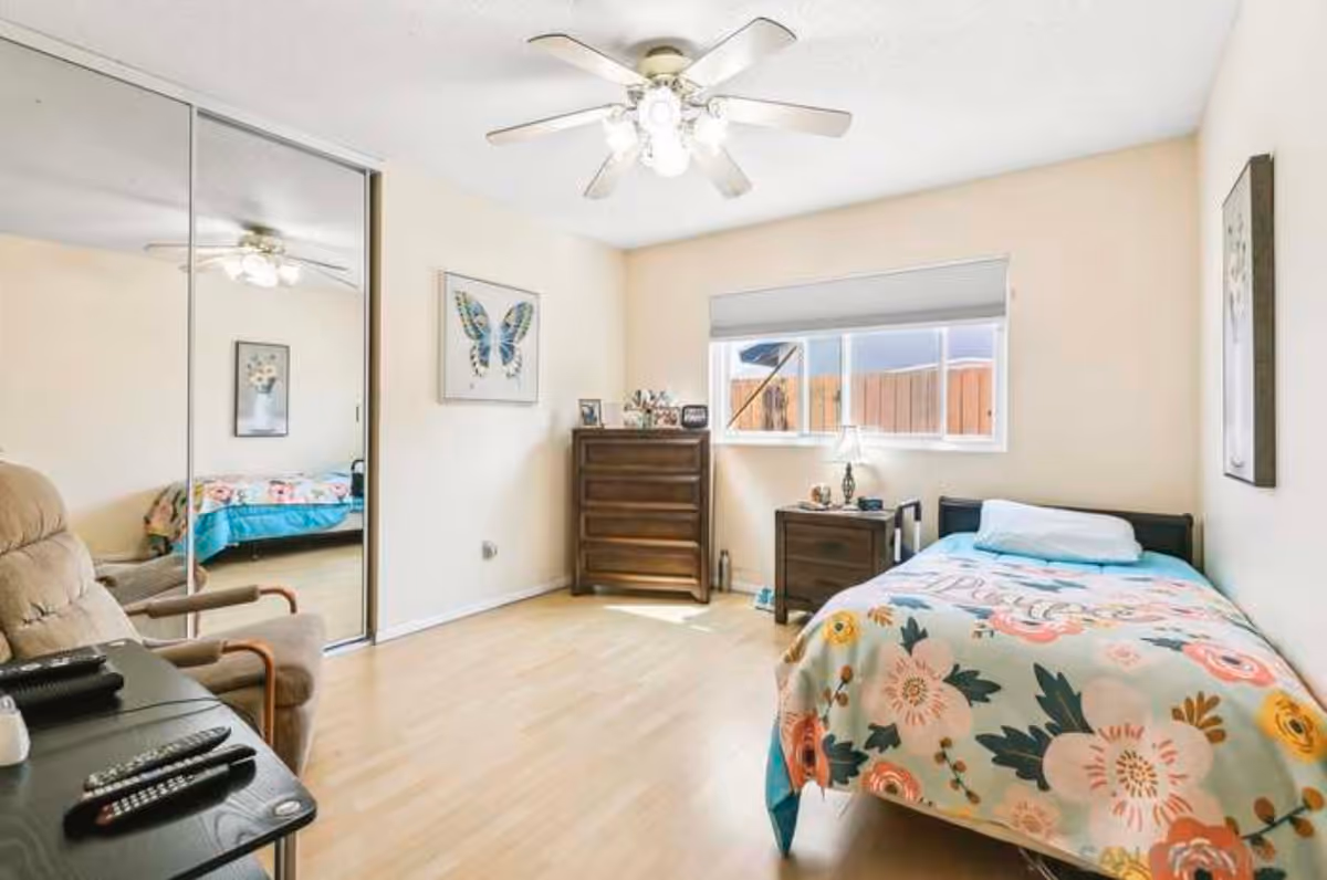A bright bedroom with light wooden flooring, a single bed with a floral patterned blanket, a wooden dresser, a nightstand with a lamp, a beige recliner chair, and a ceiling fan with lights. The room has a large window with a view of a wooden fence and mirrored closet doors reflecting part of the room.