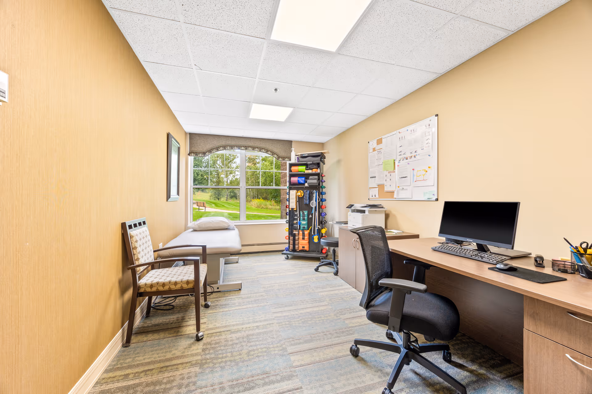 A well-lit office or therapy room with a large window showing a green outdoor area. The room contains a desk with a computer, an office chair, a printer, a bulletin board with papers pinned on the wall, a chair with armrests, and a padded examination or therapy table. There is also a rack with various exercise or therapy equipment near the window.