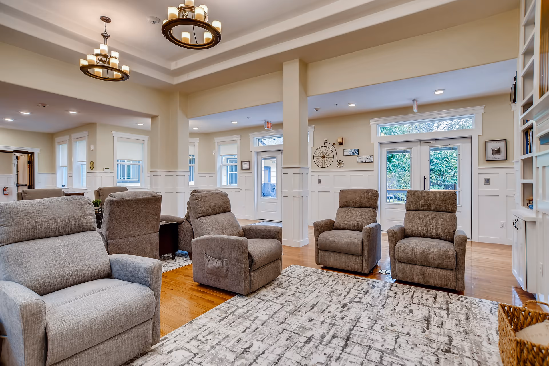 A bright and spacious living room area in a senior living facility with several comfortable gray recliner chairs arranged on a patterned rug. The room features white paneled walls, large windows, glass doors leading outside, and modern circular chandeliers hanging from the ceiling. There is a built-in bookshelf on the right side and wooden flooring throughout.