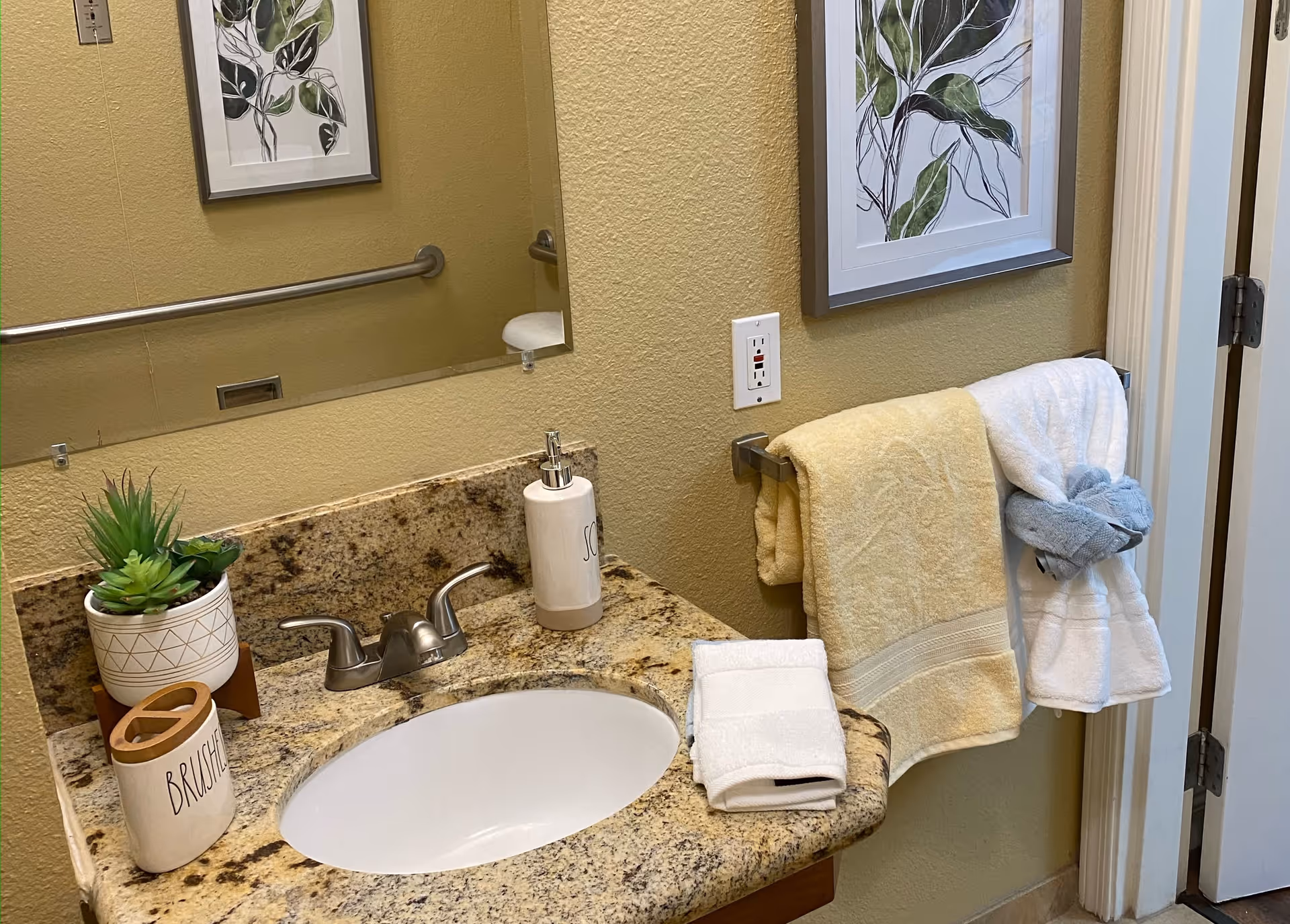 A bathroom sink area with a granite countertop, a silver faucet, a soap dispenser, a container labeled 'BRUSHES', a small potted plant, and neatly folded towels on the counter and hanging on a towel rack. A framed botanical print hangs on the wall above the towel rack.