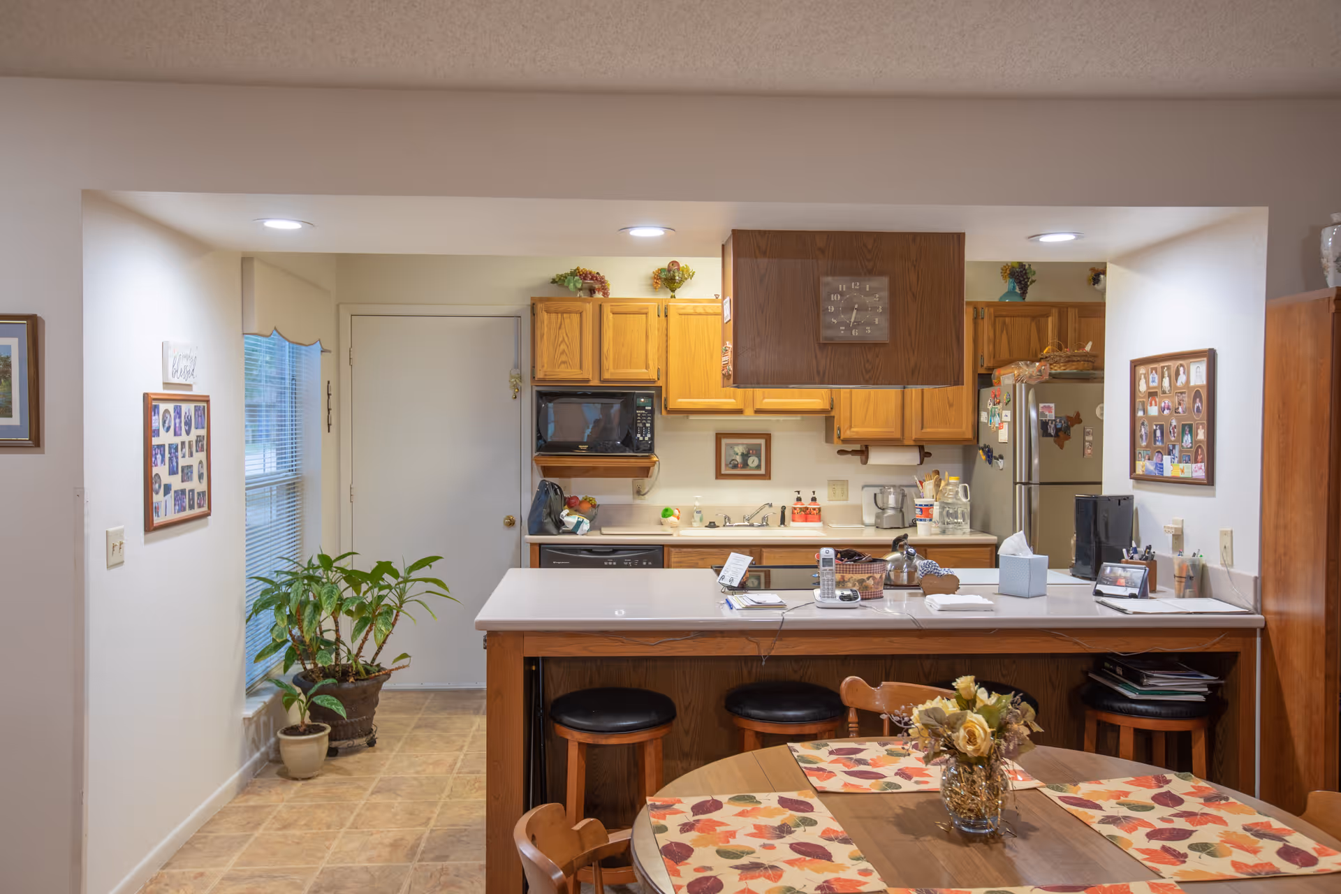 Interior view of a kitchen area with wooden cabinets, a microwave, a refrigerator covered with magnets, and a countertop with stools. In the foreground, there is a round dining table with autumn-themed placemats and a vase of flowers. The room has tiled flooring and some framed pictures on the walls.
