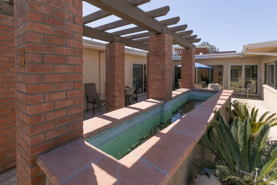 Outdoor courtyard area with a long rectangular water feature bordered by red brick columns and a wooden pergola overhead. Several chairs are placed along the building walls, and there are some green plants near the water feature. The sky is clear and blue.