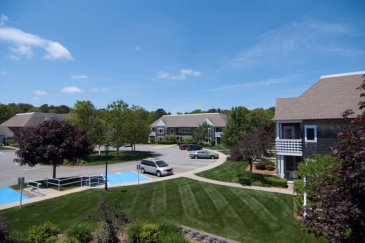 View of a senior living facility parking lot and surrounding buildings at Thirwood Place on a sunny day with blue sky, green grass, trees, and several parked cars.