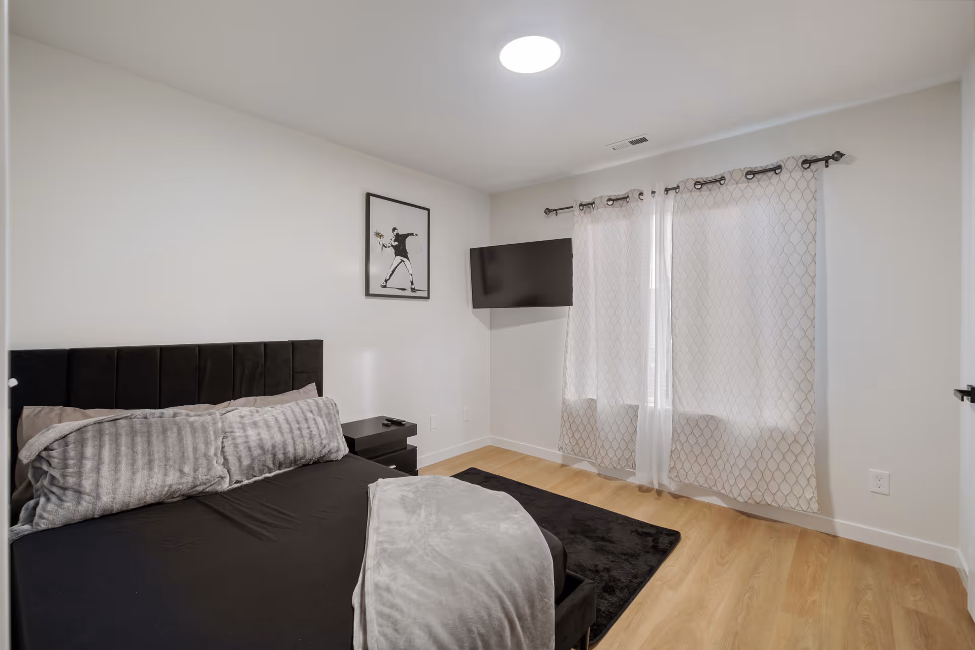 A modern bedroom with a black upholstered bed, gray pillows and blanket, a black nightstand, a wall-mounted flat screen TV, light wood flooring, and a window with patterned curtains letting in natural light. A framed black and white artwork hangs on the wall above the nightstand.