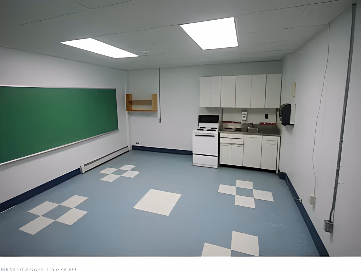 A small room with a kitchenette featuring white cabinets, a stove, and a sink. The floor has a blue and white checkered pattern, and there is a large green chalkboard on one wall. The ceiling has fluorescent lighting panels.