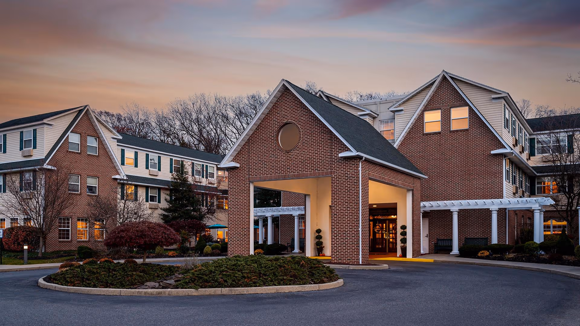Exterior view of a senior living facility named Atria Bethlehem during sunset, showing a brick building with multiple windows, a covered entrance with a peaked roof, landscaped greenery, and a circular driveway.