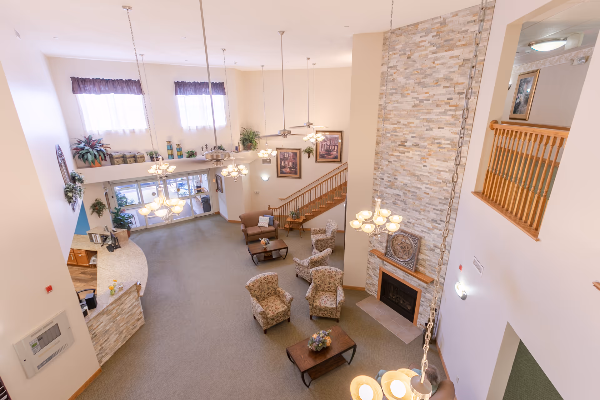A spacious and well-lit senior living facility common area with high ceilings, hanging chandeliers, a stone fireplace, several upholstered armchairs, coffee tables with floral arrangements, and a staircase leading to an upper floor. Large windows with curtains allow natural light to fill the room, and there are decorative plants and framed artwork on the walls.