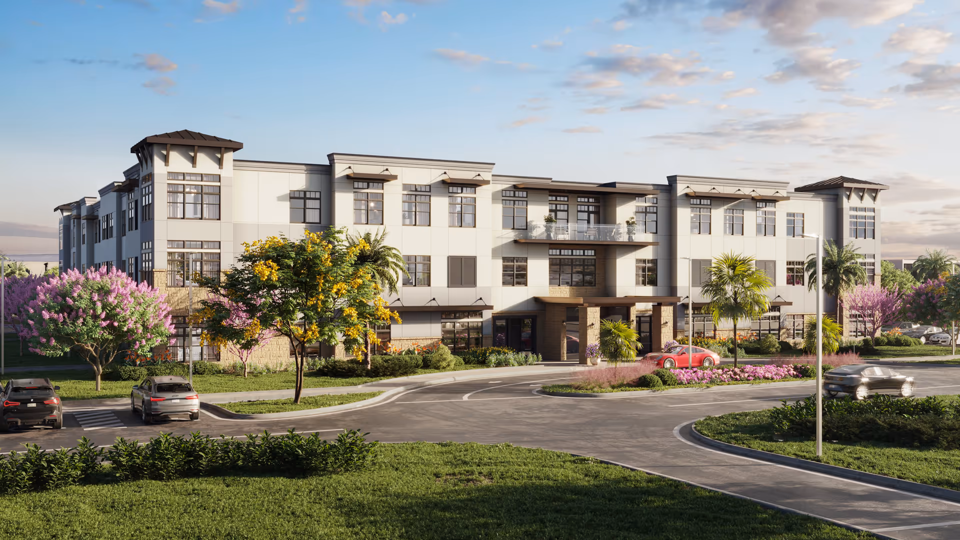 Exterior view of a modern three-story senior living facility named The Palms at Plantation, surrounded by landscaped greenery, colorful flowering trees, and parked cars under a partly cloudy sky.